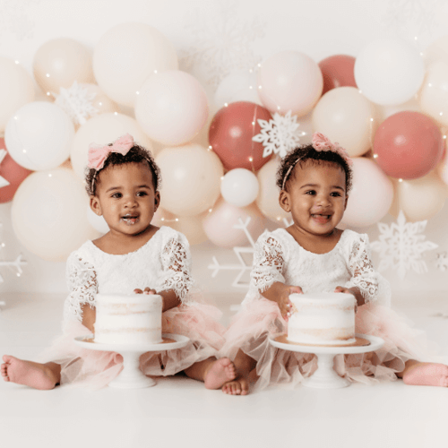 Twin girls sitting in front of balloon arch with birthday cakes