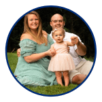 a mother wearing a green dress sits on the grass next to her husband and infant daughter