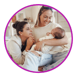 a same sex female couple holds their newborn son while sitting on their bed smiling