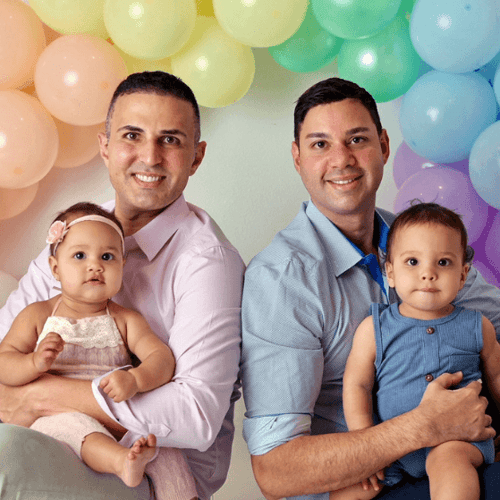 A gay couple holds their infant twins in front of a rainbow balloon arch