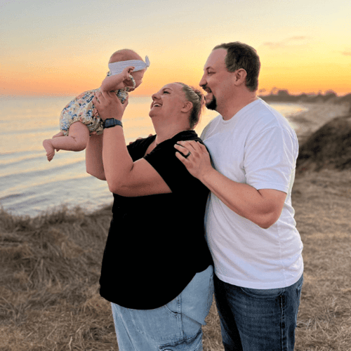 Happy new parents hold their daughter up in front of a beach sunset