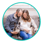 new parents smile down at their baby girl while sitting on couch with their two dogs