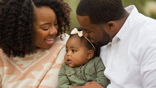 Black mother and father smiling while holding infant daughter dressed in green outfit with white bow headband