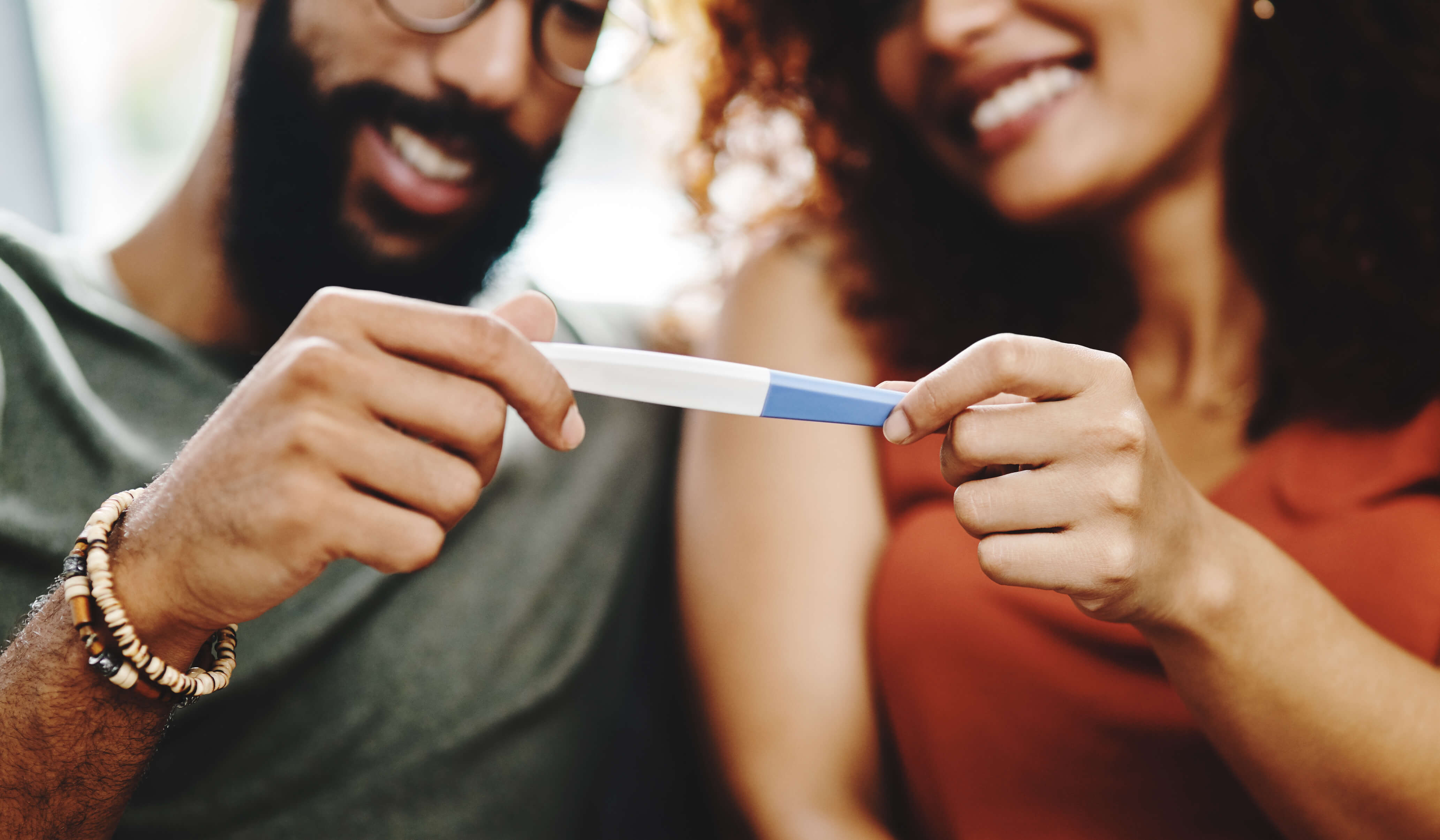 A smiling couple sits together and looks down at a positive pregnancy test they are holding between them.