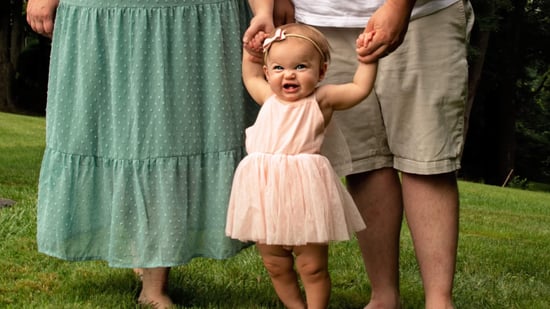 baby girl standing in front of her parent's legs wearing light pink dress