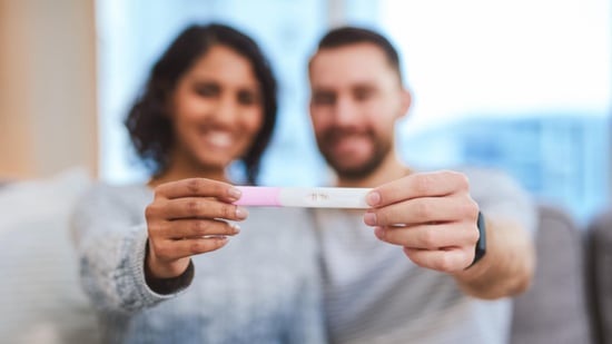 smiling male female couple blurred in background holds positive pregnancy test in foreground 