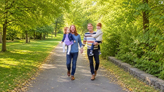 Two dads walk hand in hand down a tree-lined path on a sunny day, each carrying one of their two young children, a toddler girl in a purple dress and a young boy with red hair in a cream sweater.