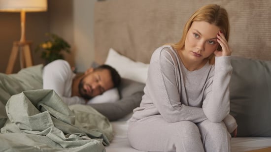 woman sits on edge of bed upset while her partner sleeps in the background 