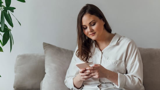 woman wearing a white shirt holds her smart phone looking down at it with a grey pillow behind her